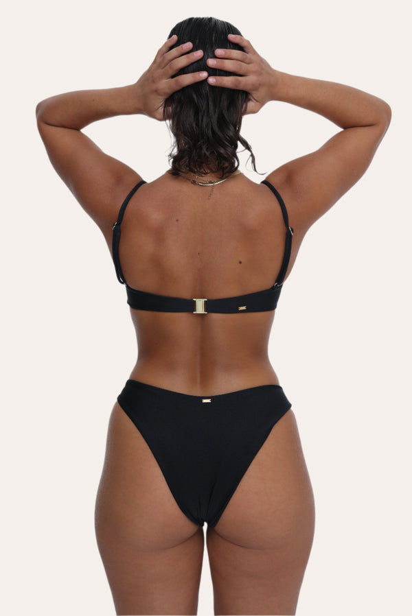 Amour black bikini Bottoms in Onyx with a gold ring, Sustainable recycled swimwear luxury bikini, seen on a model with both arms on her hair